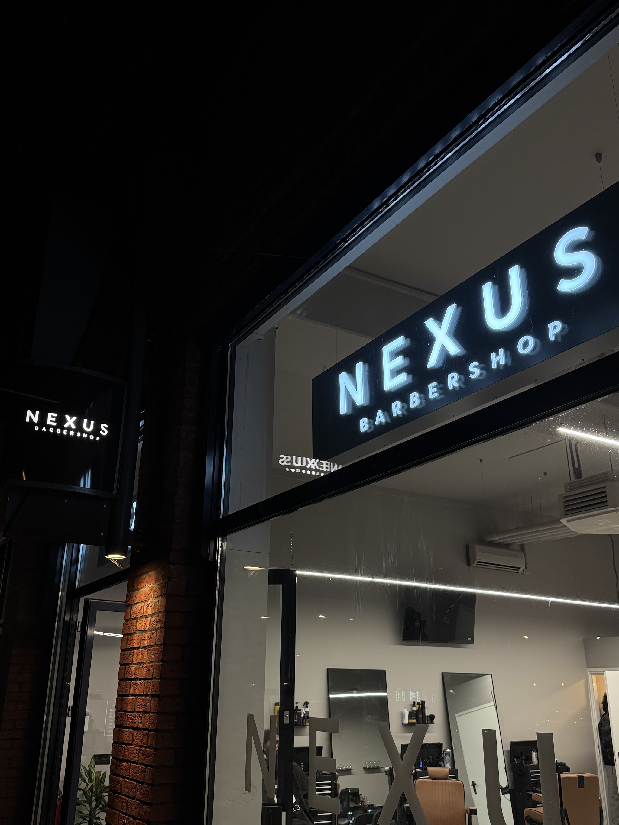 De gevel van Nexus Barbershop in het donker, met de verlichte letters NEXUS BARBERSHOP boven het raam